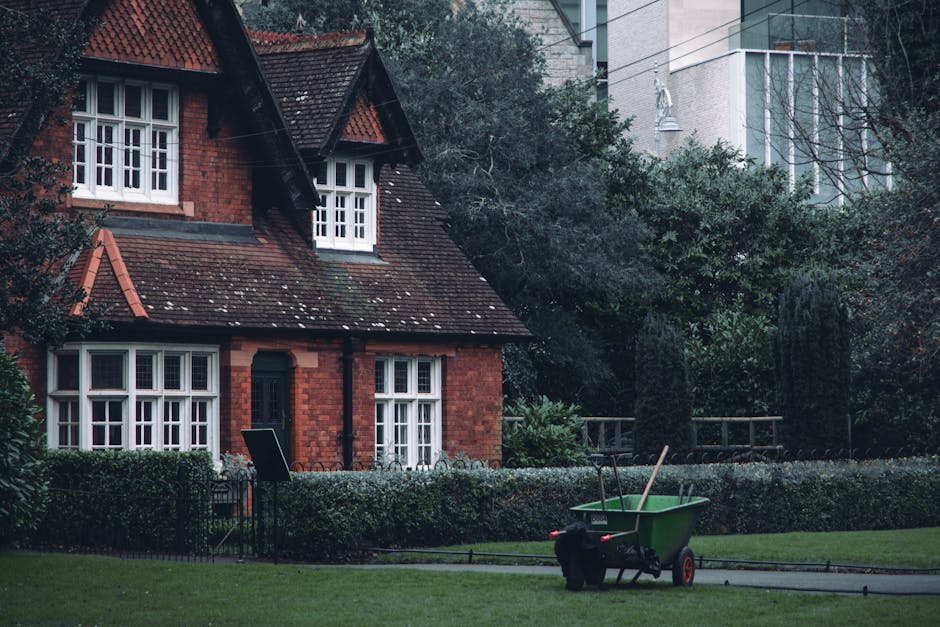 A residential garden featuring a well-maintained lawn with lush, trimmed grass bordered by a low hedge. In the foreground, a green garden wheelbarrow with a black dog sitting beside it contains two ga